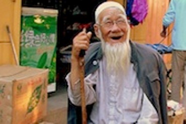 An old man, smiling with his walking stick sit on a stool