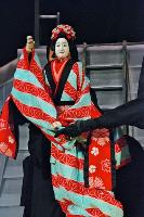A female Sanbaso Bunraku puppet wearing a red kimono being manipulated by 2 puppeteers